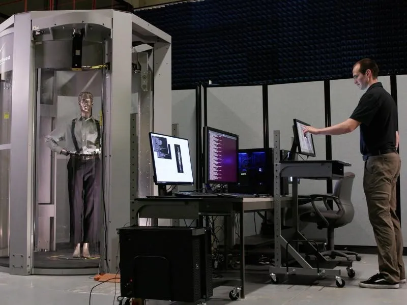 A mannequin is positioned in a stance with hands on its hips inside an airport screening machine in a laboratory setting while an individual observes Passenger Screening Retrofit Kit capabilities displayed on three computer monitors.