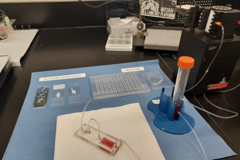 3D printed lung tissue is on a microchip within a laboratory slide that is connected via tubes to other instruments. Labels above a group of other slides reads: 3D PRINTED MICROFLUIDS. Another label above a receptacle reads: INNOVATIONS.