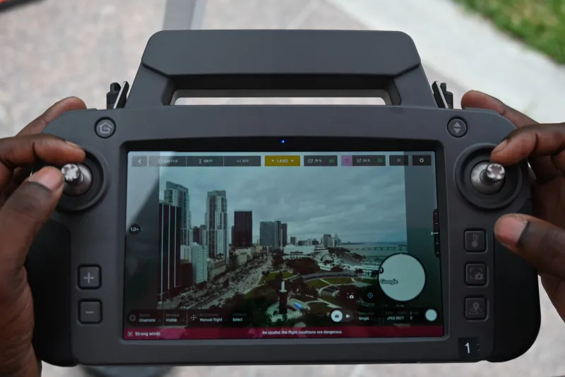 A first responder looks at a Parrot ANAFI USA GOV drone’s ground control station (GCS) while conducting a flyover of Bayfront Park. The GCS is large, rectangular, and black; and has a variety of controls and buttons that are used to fly/navigate the drone and utilize its various capabilities. The display screen on the GCS shows an aerial view of the park.