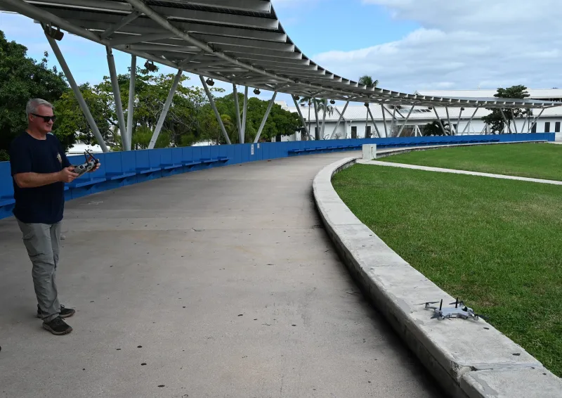 A Teal 2 drone sits on a sidewalk in the Bayfront Park Amphitheater. A first responder stands nearby as he calibrates the drone and prepares it for launch. In the background: an awning and blue amphitheater seats surround the first responder and the drone.