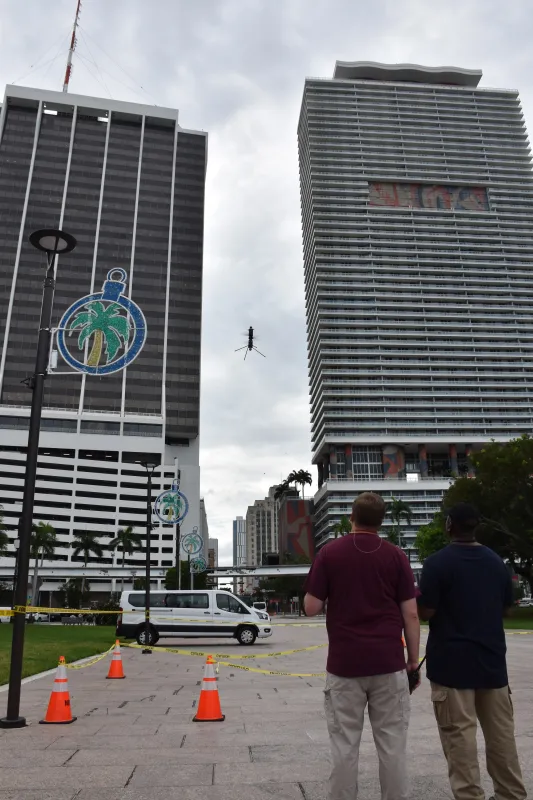 An Ascent AeroSystems Sprit drone prepares to land in Bayfront park after conducting a damage assessment scenario. An MSU SME (standing on the left) observes as an evaluator (standing on the right) guides the drone to the ground with its GCS. In the background: skyscrapers and the skyline surrounding Bayfront park.