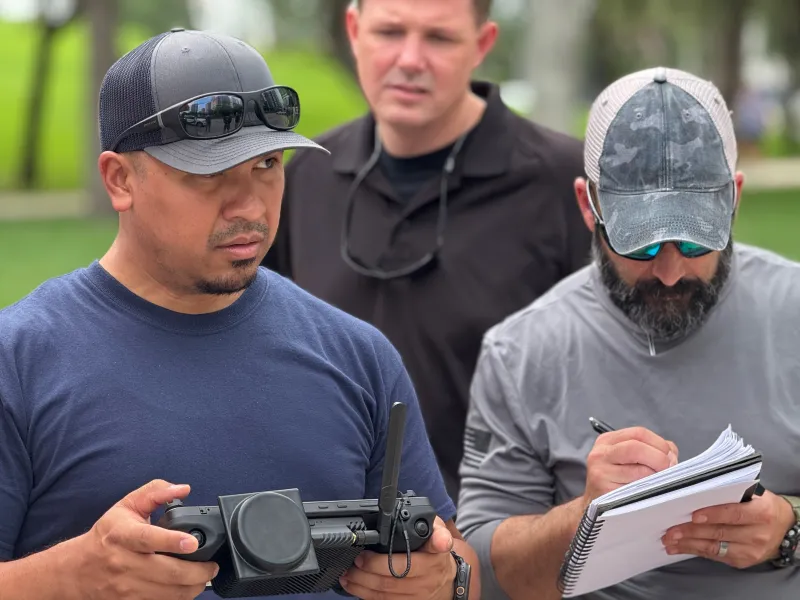 A first responder uses a GCS to fly an Ascent AeroSystems Spirit drone during a scenario-based flight. The first responder standing on the left and in the foreground is controlling the drone with the GCS,  which is black and rectangular. To his right stands a DAGER Technology data collector who taking notes based on his operational feedback. 