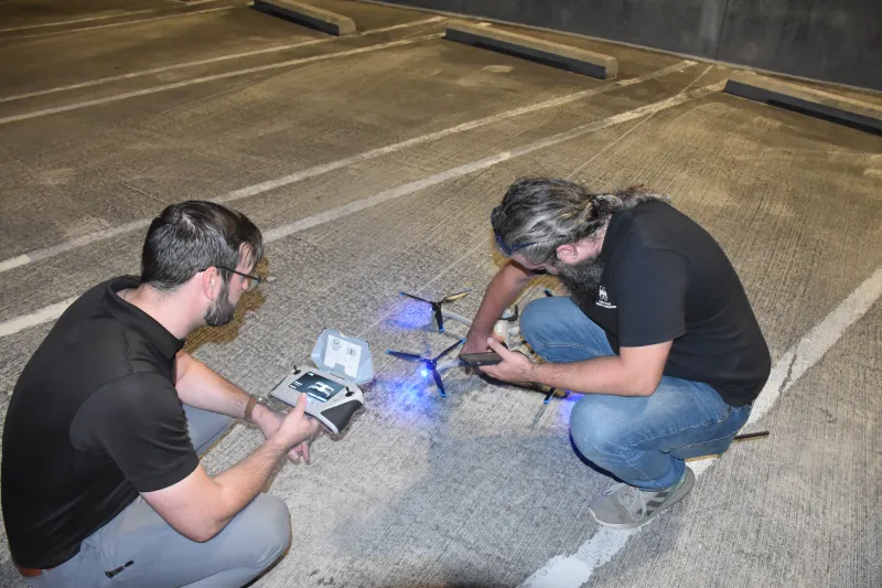 A responder and MSU subject matter expert both kneeling over a Skydio X10D drone and sync it with its GCS in preparation for a launch. The drone sits on a concrete parking lot and has its headlights on. 