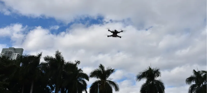A black Teal 2 drone soars above the trees of Bayfront Park in Miami, Florida. Four upward-facing propellors help it fly over the park. In the background: tall trees, a bright blue sky, and white clouds stand out in stark contrast to the drone.