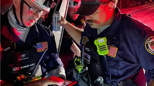 Two US&R Florida Task Force 2 (FL-TF2) personnel, dressed in blue tactical uniforms, are using SARCOP on a cell phone. It is nighttime and they are standing close to a vehicle with a third FL-TF2 member in it. There appears to be white sand on the road.