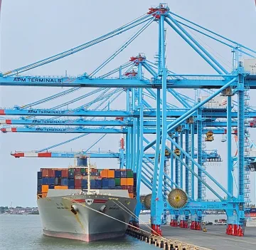 A white ship, seen from the front, awaits inspection floating in calm water at the Miami Seaport. The ship, tied with ropes to the pier, is loaded with colorful shipping containers. On the pier, multiple turquoise ship-to-shore cranes stand, ready to load or unload the ship. A light blue sky and cityscape is in the background.